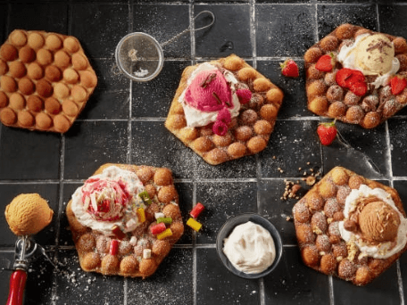 Various bubble waffles are topped with ice cream, strawberries, whipped cream, and colorful candies