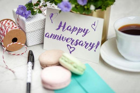 A "Happy anniversary!" card is surrounded by macarons, a cup of tea, a pen, and a spool of red and white twine