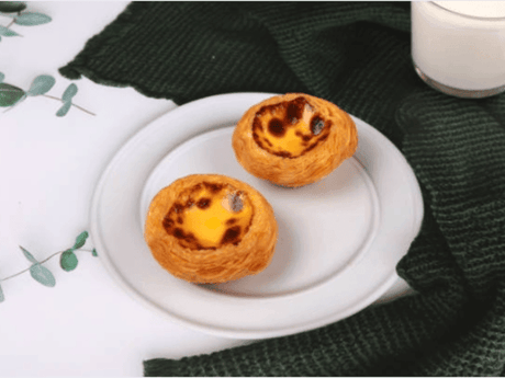 Two Portuguese egg tarts sit on a white plate next to a glass of milk and a green cloth.