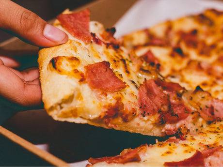 Hand holding a slice of pizza with melted cheese and ham from a pizza box