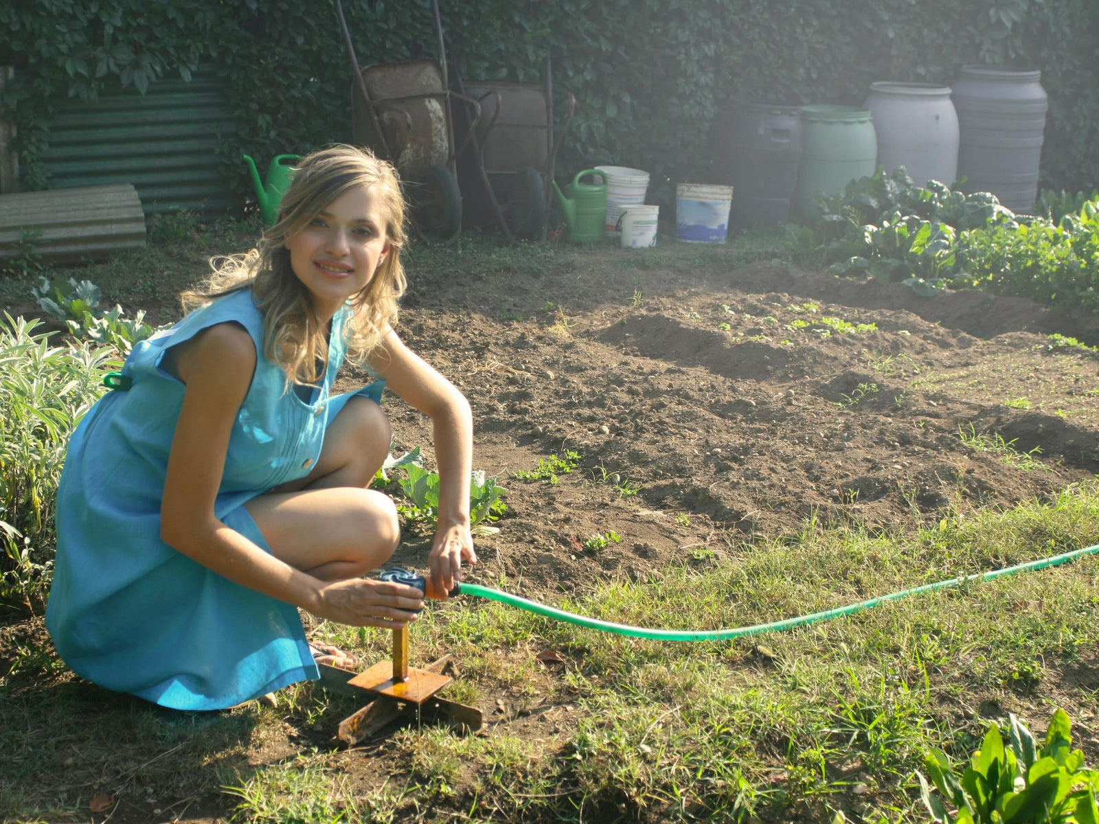 A woman wearing a turquoise dress tending to her vegetable garden using a garden hose and sprinkler setup