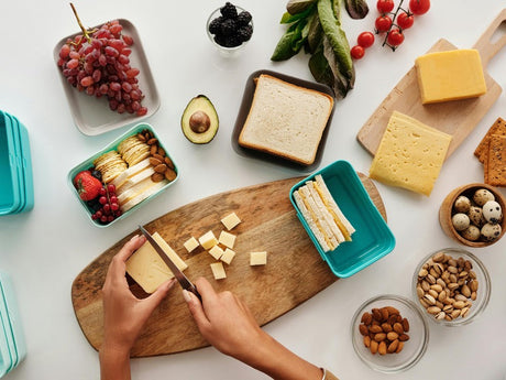 a person cutting cheese on a wooden board, surrounded by various snacks including grapes, crackers, nuts, and sandwich ingredients