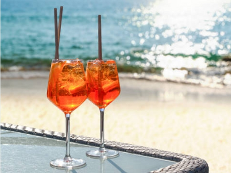 Two glasses of Aperol Spritz cocktails with ice and straws are placed on a table by the beach