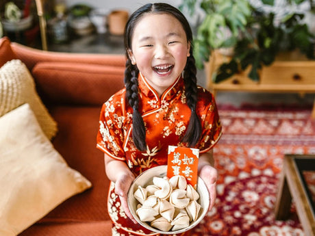 A young girl in traditional Chinese attire is smiling while holding a bowl of dumplings and a red envelope