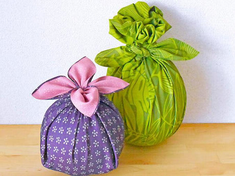 Two fabric-wrapped round gifts, one purple with a pink bow and the other green with a leaf pattern, sit on a wooden surface