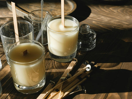 Two homemade candles in glasses sit on a wooden table next to crafting tools and materials