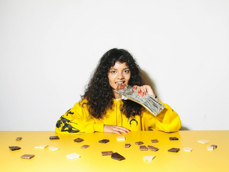 A woman in a yellow hoodie enjoying a chocolate bar, surrounded by scattered chocolate pieces