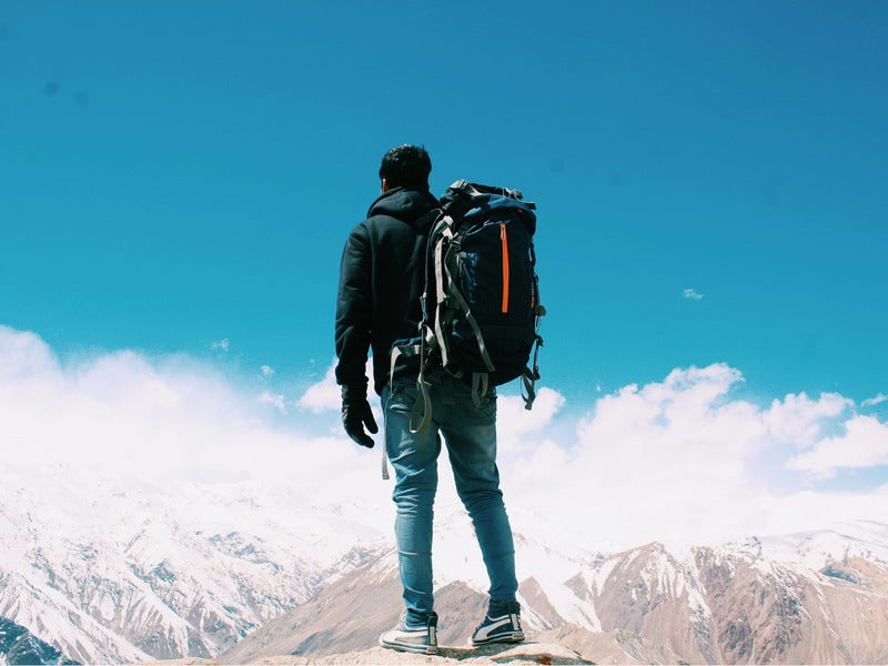 A hiker with a backpack stands on a rocky outcrop, overlooking snow-capped mountains under a clear blue sky