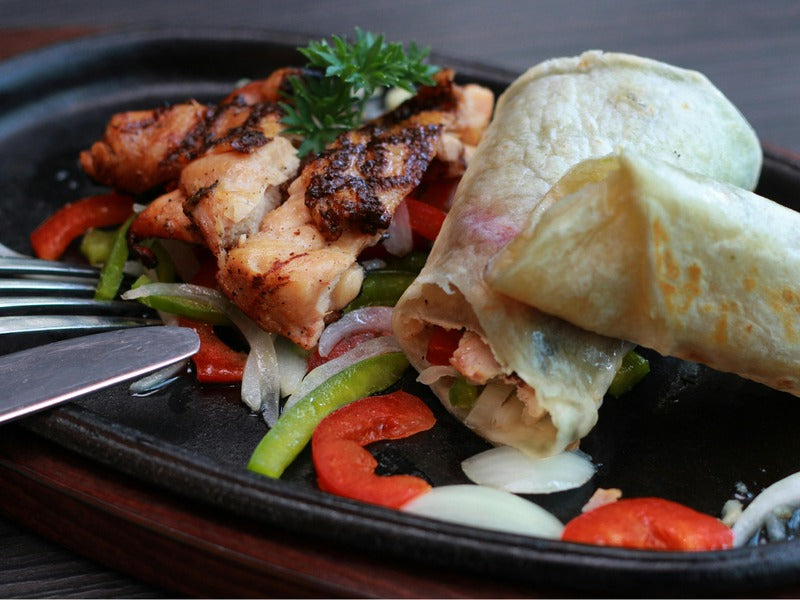 A sizzling plate features grilled chicken, bell peppers, onions, and a tortilla wrap, garnished with parsley