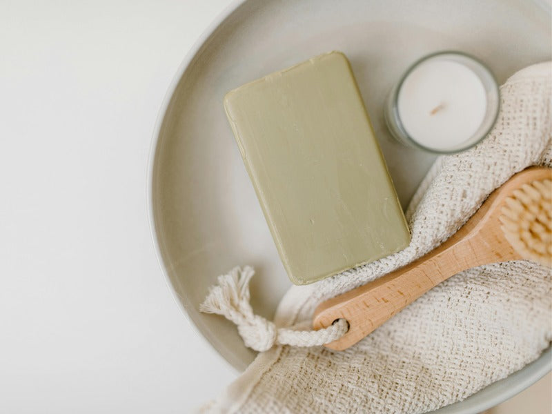 a minimalist self-care setup featuring a natural soap bar, a wooden body brush, an exfoliating mitt, and a candle