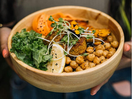 A wooden bowl filled with a healthy meal including kale, chickpeas, roasted vegetables, and a lemon wedge