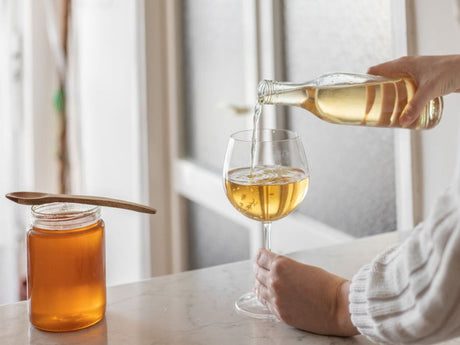 A person pouring homemade mead into a wine glass beside a jar of honey