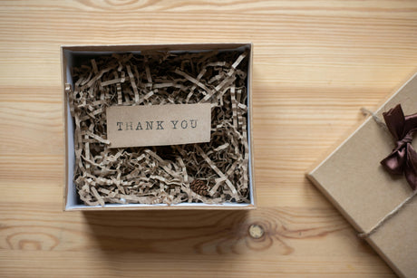 Open gift box filled with brown crinkle paper and a “Thank You” card on top